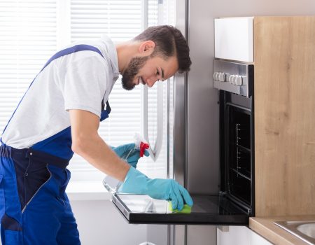 Happy Male Janitor Cleaning Oven With Sponge In Kitchen